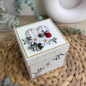 Op de afbeelding: Een witte doos met een hartvormig ontwerp aan de voorkant. Het hart is gemaakt van hout met een witgewitte afwerking en heeft een gouden rand. De doos is versierd met witte bloemen, groene bladeren en een rode lieveheersbeestje. De doos staat op een geweven rieten oppervlak.