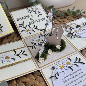 Op de afbeelding: Een witte en gouden trouwkaart met een pop-up ontwerp met een stel dat voor een bloemstuk staat. De kaart is versierd met witte madeliefjes en groene bladeren. De tekst op de kaart luidt "Sandra & Bo&scaron;tjan".