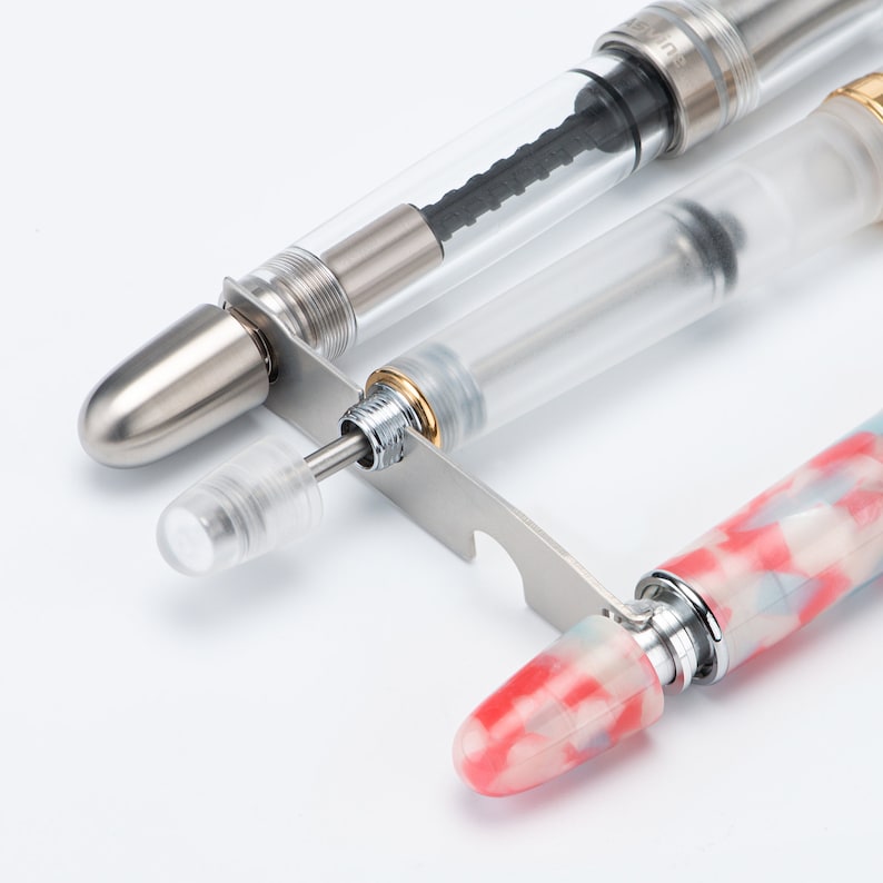 May include: A close-up of three fountain pens with different filling mechanisms. The pens are on a white surface. The pen on the left has a silver cap and a clear barrel. The pen in the middle has a clear barrel and a silver cap. The pen on the right has a red and white marbled barrel and a silver cap.