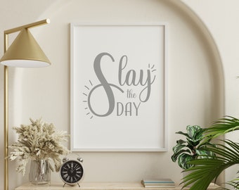 May include: A white framed print with the text "Slay the Day" in gray cursive lettering. The print is hanging on a white wall above a wooden shelf with a gold lamp, a vase with dried flowers, a silver alarm clock, and a stack of books.