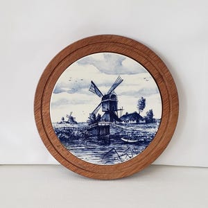 May include: Round wooden framed decorative plate featuring a blue and white illustration of windmills on a river. The artwork depicts a rural landscape with windmills, water, and a cloudy sky. The plate is a decorative item.