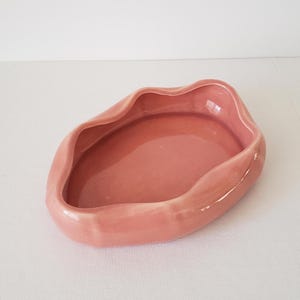 May include: A pink ceramic dish with an organic, wavy shape. The dish has a smooth, glossy finish and a shallow interior. The color is a muted, dusty rose. The dish is likely for decorative or functional use.