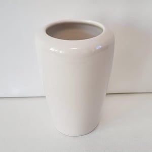 May include: A cream-colored ceramic vase with a glossy finish. The vase has a cylindrical shape with a slightly wider top opening. The interior of the vase is a lighter shade of cream. This decorative vase is suitable for floral arrangements.