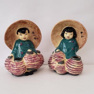 May include: Pair of ceramic figurines depicting Asian figures. Each figure wears a green tunic, a straw hat, and carries multiple lanterns. The lanterns are primarily pink with a striped design. The figurines have black hair and are set against a white background.