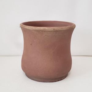 May include: A terracotta-colored, handmade ceramic pot with a rounded base and flared rim. The pot has a smooth, unglazed surface and a slightly uneven texture, suggesting a rustic aesthetic. Ideal for indoor or outdoor plants.