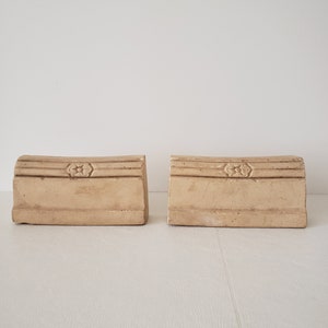 May include: Two beige ceramic bookends with a floral design. Each bookend has a raised floral design on the front and a slightly curved top.