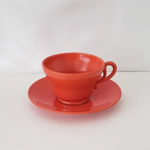 May include: A vibrant red teacup and saucer set. The cup has a rounded shape with a handle, and it sits on a matching saucer. The set is made of ceramic or porcelain and is suitable for serving tea or coffee. The set is a solid, bright red color.