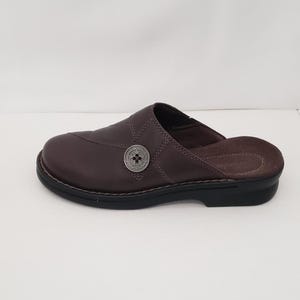 May include: Brown leather clog-style shoe with a black sole. The shoe features a decorative button on the side and stitched detailing. The open-back design offers easy wear. A comfortable and stylish footwear option.