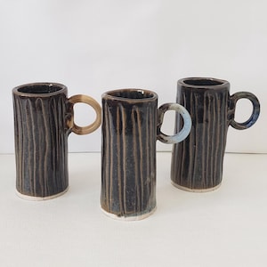 May include: Three tall, cylindrical ceramic mugs with a dark brown, vertically ribbed texture and a glossy finish. Each mug features a small, circular handle in a contrasting color: one beige, one light blue, and one dark blue, with white bases.