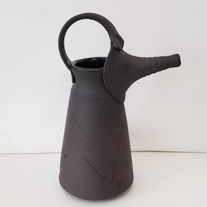 May include: A matte black ceramic pitcher with a unique spout and handle design. The pitcher has a conical shape with a stitched pattern detail. The handle is a rounded arch. The spout extends horizontally from the body.