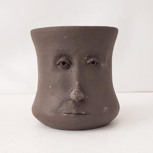 May include: A brown ceramic face planter with a human face sculpted on the front. The planter has a wide, rounded base and a flared top. The face features eyes, a nose, and a mouth. The planter is suitable for indoor or outdoor use.