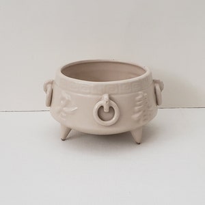 May include: A cream-colored ceramic planter with three legs and two handles. The planter has a ring-shaped design on the front and a raised design on the sides.
