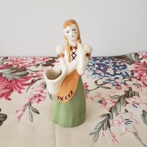 May include: A ceramic figurine of a woman with brown hair, wearing a white blouse, brown vest, and green skirt. She is holding a small white basket in her left hand. The figurine is painted with brown details, including stitching on the skirt.