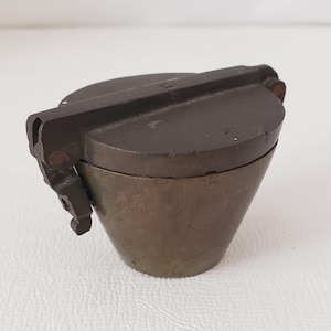 May include: A vintage, bronze-colored metal container with a conical base and a hinged, flat lid. The lid has a rectangular handle and a small, decorative clasp. The container is likely used for storage or display.