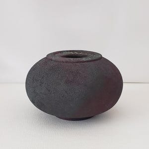May include: A round, dark-colored ceramic vase with a textured surface. The vase has a wide, open top and a slightly bulbous shape. The color transitions from dark gray to a reddish-brown hue.