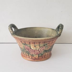 May include: A small, brown ceramic planter with two green rope handles. The planter is decorated with a basket weave pattern and a floral design featuring pink roses and green leaves.