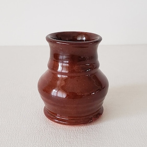 May include: A small, brown ceramic vase with a rounded body and a flared rim. The vase has a glossy finish and is perfect for displaying small flowers or greenery.