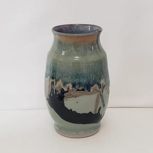 May include: A ceramic vase with a flared rim and a rounded body. The glaze features a blend of sage green, deep blue, and black tones, with a distinct drip pattern cascading down the sides. The interior rim shows a dark purple and brown hue.