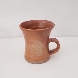 May include: A terracotta-colored ceramic mug with a speckled finish and a flared rim. The mug features a smooth, glossy glaze with a subtle, lighter-toned abstract swirl pattern on the front and a sturdy, curved handle on the right side.
