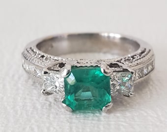 May include: A silver ring featuring a large, square-cut emerald center stone flanked by two smaller, square-cut diamonds. The band has a detailed, textured design. A classic piece of jewelry.