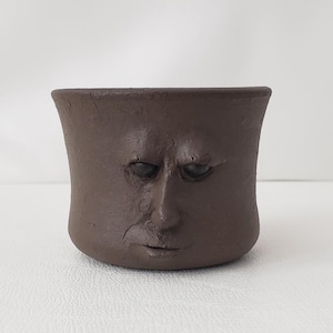 May include: A brown ceramic planter with a sculpted face. The planter has a wide opening and a slightly flared rim. The face has a furrowed brow and a contemplative expression. The planter is suitable for small plants.