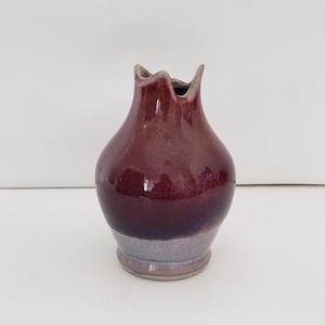 May include: A small, handcrafted ceramic vase with a deep burgundy glaze. The vase has a bulbous body and a flared, scalloped rim. The base is a lighter shade of purple. The vase is approximately 5 inches tall.