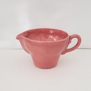 May include: A pink ceramic creamer with a handle and a spout. The creamer has a rounded body and a flared rim. The handle is a loop shape. The creamer is likely vintage or antique.