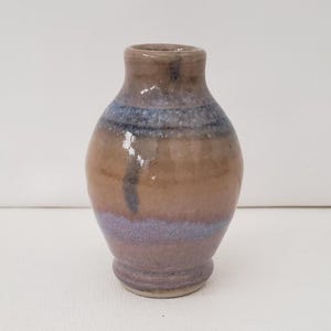 May include: A small, handcrafted ceramic vase with a rounded body and a narrow neck. The vase features a color gradient, transitioning from a light purple base to a brown midsection, and a blue-grey top. The vase is likely used for decorative purposes.