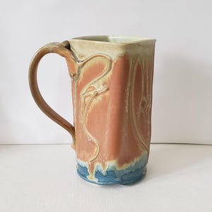 May include: Handmade ceramic mug with a square top and a curved handle. The mug features a textured design with a coral-colored body, cream-colored top, and blue base. The mug is approximately 6 inches tall.