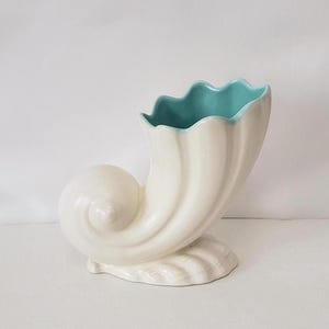 May include: A white ceramic vase in the shape of a conch shell. The interior of the vase is a light teal color, and the rim has a scalloped edge. The vase has a spiral design and sits on a base with a shell-like texture.