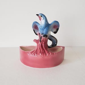 May include: A pink ceramic planter shaped like a bird perched on a tree branch. The bird is blue with a pink breast and black wings.