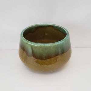 May include: A handcrafted ceramic bowl with a rounded base and a wide opening. The bowl features a two-tone glaze, with a green upper half and a brown lower half. The interior is a darker brown. The bowl is suitable for serving or display.