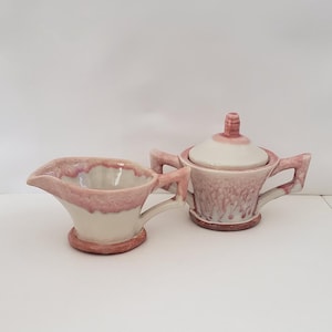May include: A cream-colored ceramic creamer and sugar bowl set with a pink glaze. The creamer has a spout and handle, while the sugar bowl has a lid and handles. The set is likely vintage and adds a touch of elegance to any table setting.
