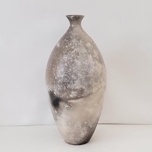 May include: A tall, teardrop-shaped ceramic vase featuring a mottled, smoky finish in shades of charcoal gray, slate, and off-white. The surface displays a cloudy, atmospheric pattern with dark patches and lighter, speckled areas, tapering to a narrow neck.