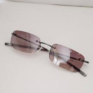 May include: Rimless sunglasses with rectangular, gradient brown lenses and a thin, dark metal frame. The sunglasses have a minimalist design, suitable for everyday wear. The lenses transition from a darker shade at the top to a lighter shade at the bottom.