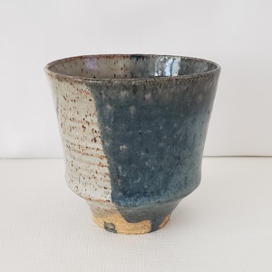 May include: Handmade ceramic cup with a unique design. The cup features a two-tone color scheme, with a speckled beige and a deep blue-gray glaze. The cup has a wide opening and a slightly flared base, perfect for beverages.
