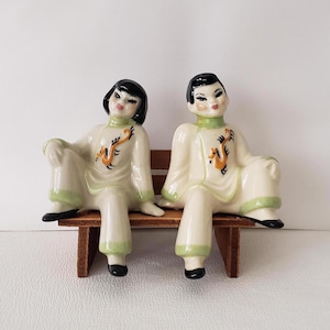 May include: A pair of ceramic figurines of people in traditional Chinese clothing sitting on a wooden bench. The figurines are painted in shades of white, green, and black, with orange accents. The figures have black hair and are wearing green and white robes with orange dragons on the front.