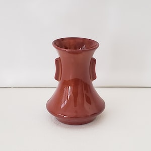 May include: A small, brown ceramic vase with two handles on the sides. The vase has a rounded bottom and a flared top.