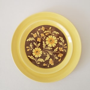 May include: A yellow ceramic plate with a brown center featuring a floral design. The flowers are yellow and orange, with brown leaves and stems. The plate has a wide yellow rim.