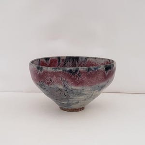 May include: A handmade ceramic bowl with a wide, slightly flared rim. The bowl features a color palette of deep red, black, and gray, with a textured, mottled appearance. The bowl's exterior has a smooth, glazed finish.