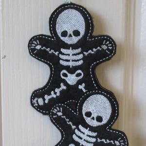 May include: Three black felt skeleton gingerbread ornaments with white embroidery. The ornaments are hanging from a white door. The ornaments are stacked on top of each other. The bottom ornament is partially obscured by the middle ornament. The top ornament is fully visible. The ornaments are decorated with white embroidery of a skeleton with a skull. The ornaments are labeled "Sew Sarah Designs" and "Machine Embroidery Designs".