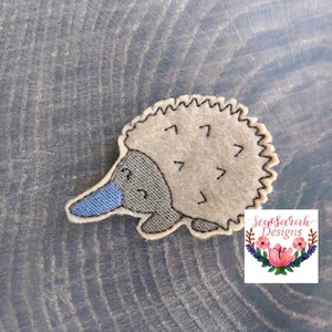 May include: A brown felt echidna applique with a blue nose and black eyes. The applique is on a brown wooden background. SewSarah Designs is printed in the bottom right corner.