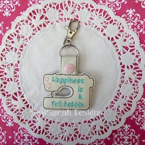 May include: White fabric keychain with a pink button and an embroidered sewing machine design. The text on the keychain reads "Happiness is a full bobbin".