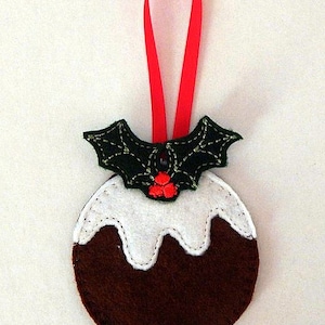 May include: A felt Christmas ornament shaped like a Christmas pudding with a green holly sprig on top. The pudding is brown with white icing and red berries. The ornament has a red ribbon for hanging.