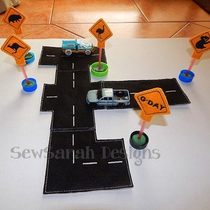 Puede incluir: Un conjunto de juego de fieltro para niños que incluye un camino negro con líneas blancas, dos coches de juguete y cinco carteles de fieltro con animales australianos y el texto "G'DAY".