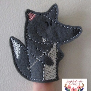 May include: Grey felt finger puppet of a wolf with a pink nose and white accents. The puppet is on a finger and the text "Sew Sarah Designs" is visible on the puppet.