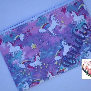 Peut inclure: Une pochette zippée violette, rose et bleue avec un motif de licorne blanche et d'arc-en-ciel. La pochette porte le nom "Bridie" brodé en violet.