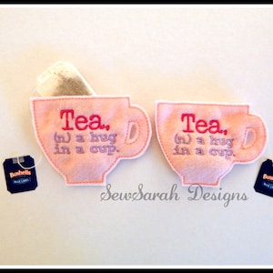 Puede incluir: Dos bolsitas de fieltro rosa en forma de taza de té con texto bordado que dice "Tea. (n) a hug in a cup." Cada bolsita contiene una bolsita de té. Las bolsitas están sobre una superficie blanca.