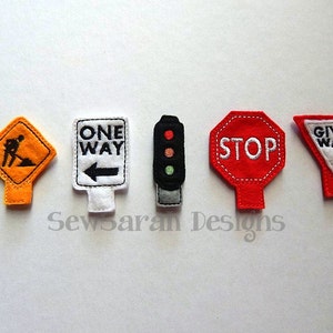 May include: Five felt road signs with white text and black outlines. The signs are:  'One Way' with a left arrow, 'Stop', 'Give Way', 'Men Working' with a stick figure, and a traffic light with red, yellow, and green lights.