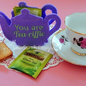 May include: Purple felt teapot tea bag holder with the text "You are Tea-riffic" embroidered on the side. A tea bag is visible inside the holder. A white teacup and saucer with a floral design are on the right side of the image. A white doily and two cookies are on the left side of the image.